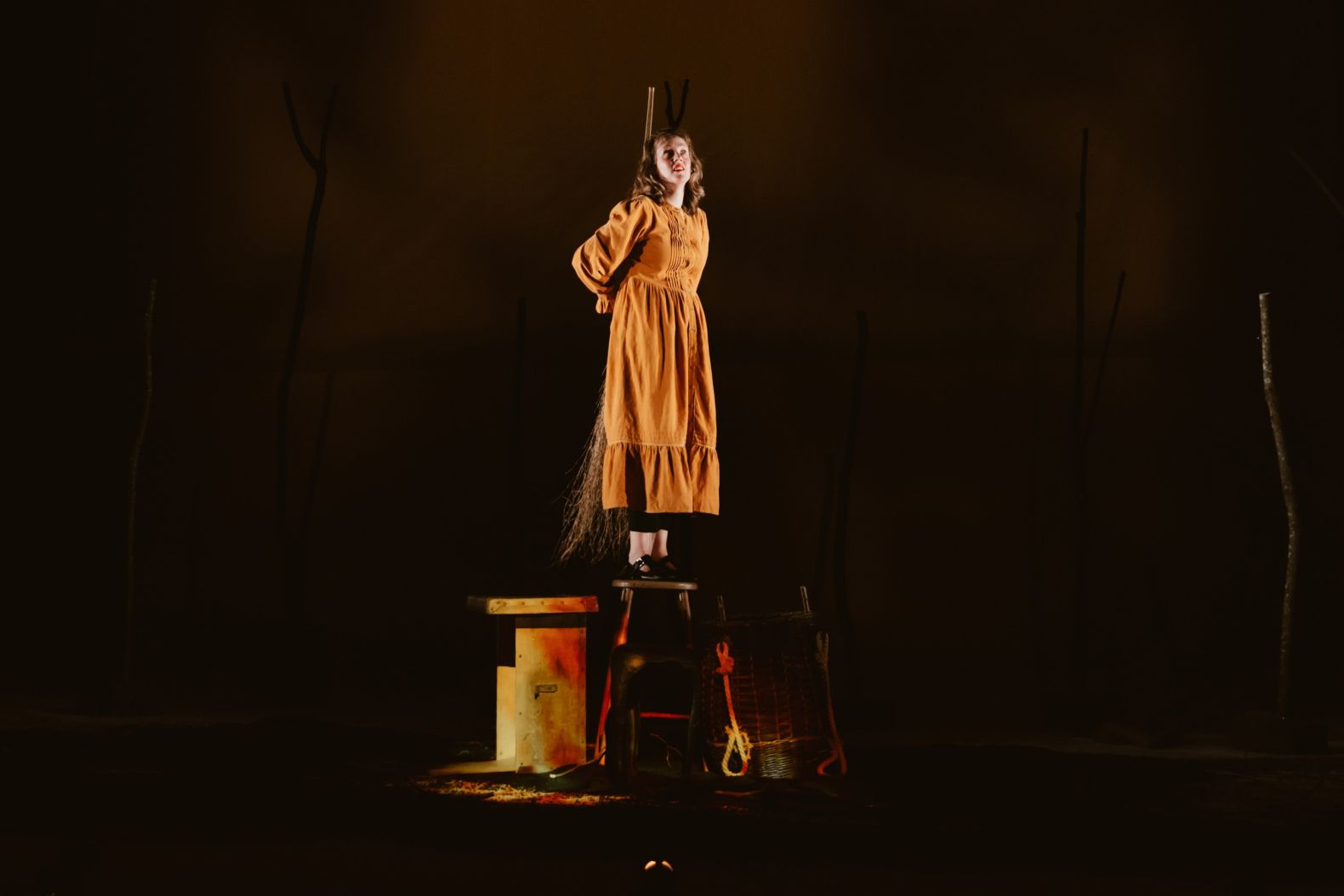 Bryony Kimmings performing in Bog Witch. Bryony is standing on a stool with a witch's broom behind her back, mimicking standing on a pire.