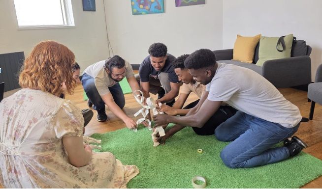 Four people kneeling around a green artificial grass mat. The group includes individuals of diverse backgrounds 