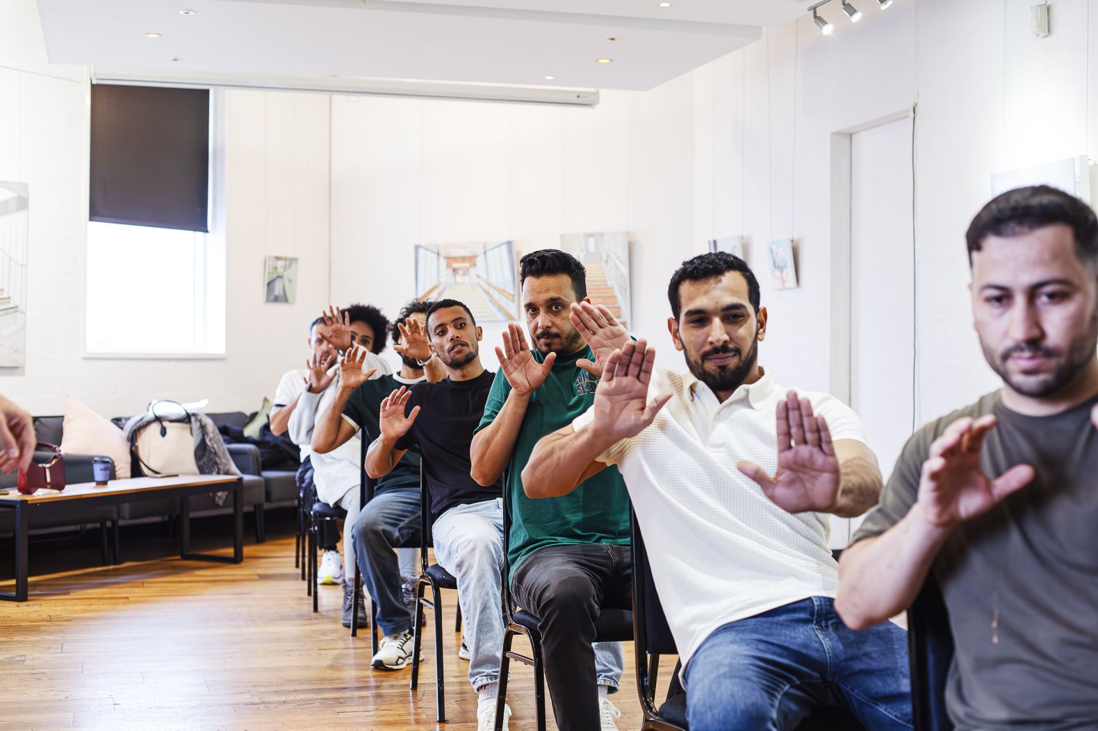 Group of men sitting in chairs in a line, raising their hands in a synchronized movement during a workshop or training session in a bright room