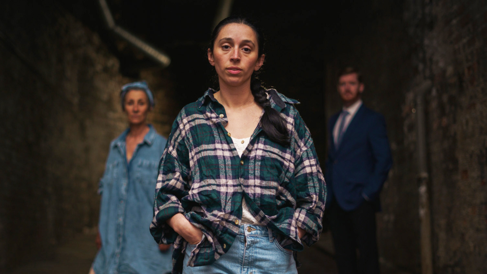A girl in her twenties, with dark brown hair that is in a low plait. She is wearing a green and pink chequered shirt and jeans.
