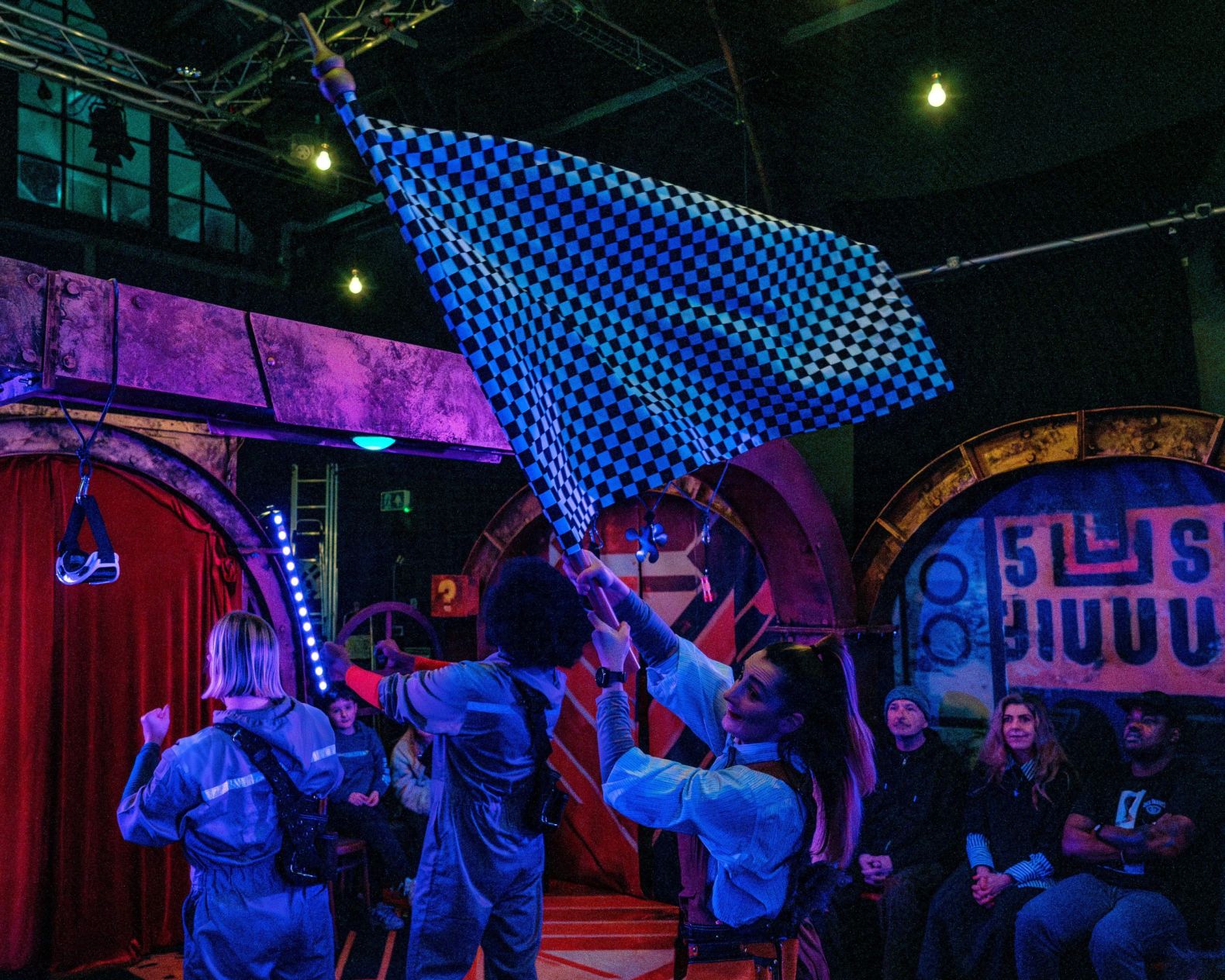 3 actors on the stage with their backs to the camera. one of them is waving a large , checked flag