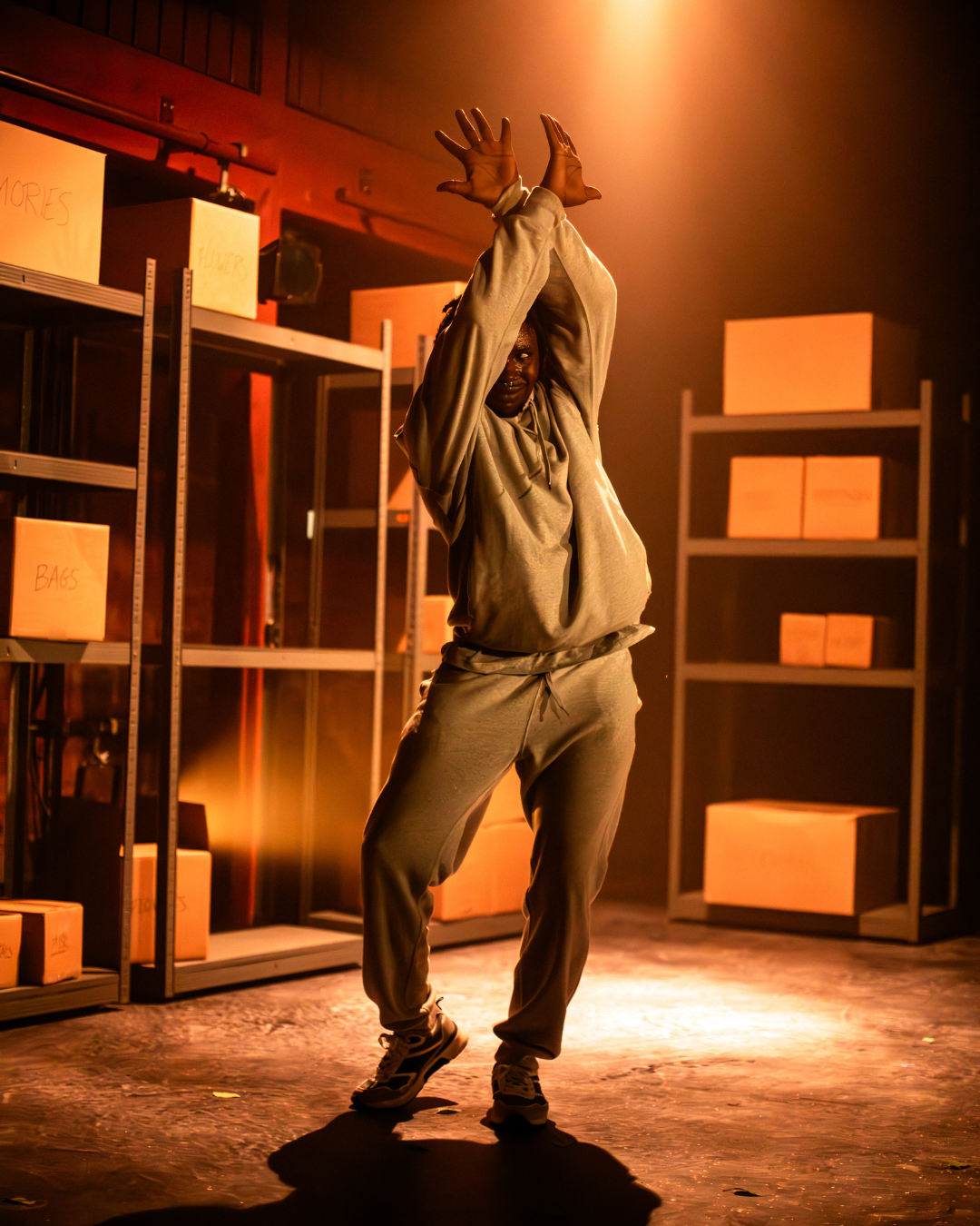 A performer is on-stage surrounded by shelves with cardboard boxes on them. The performer is dancing with his hands crossed above his head.