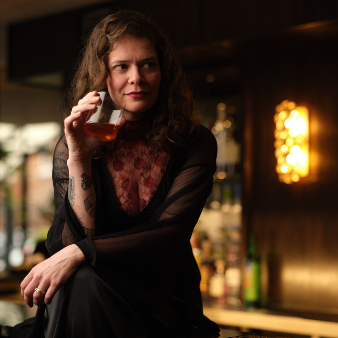 Amanda played by Hannah Ellis Ryan: she is sitting on top of the bar, wearing a black sheer dress, with red flowers and a glass of whiskey in hand.