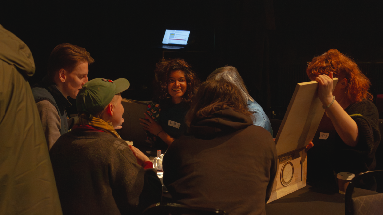 Members of Queer by Gum sitting around the table. One member is holding the lid of the wooden box that is in front of them. 