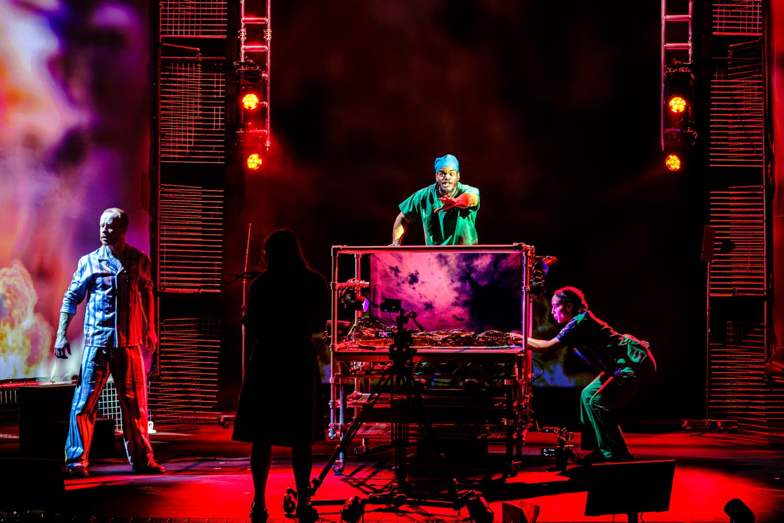Four actors on stage, surrounding a table on wheels which has a screen attached showing purple skies and a model landscape. 