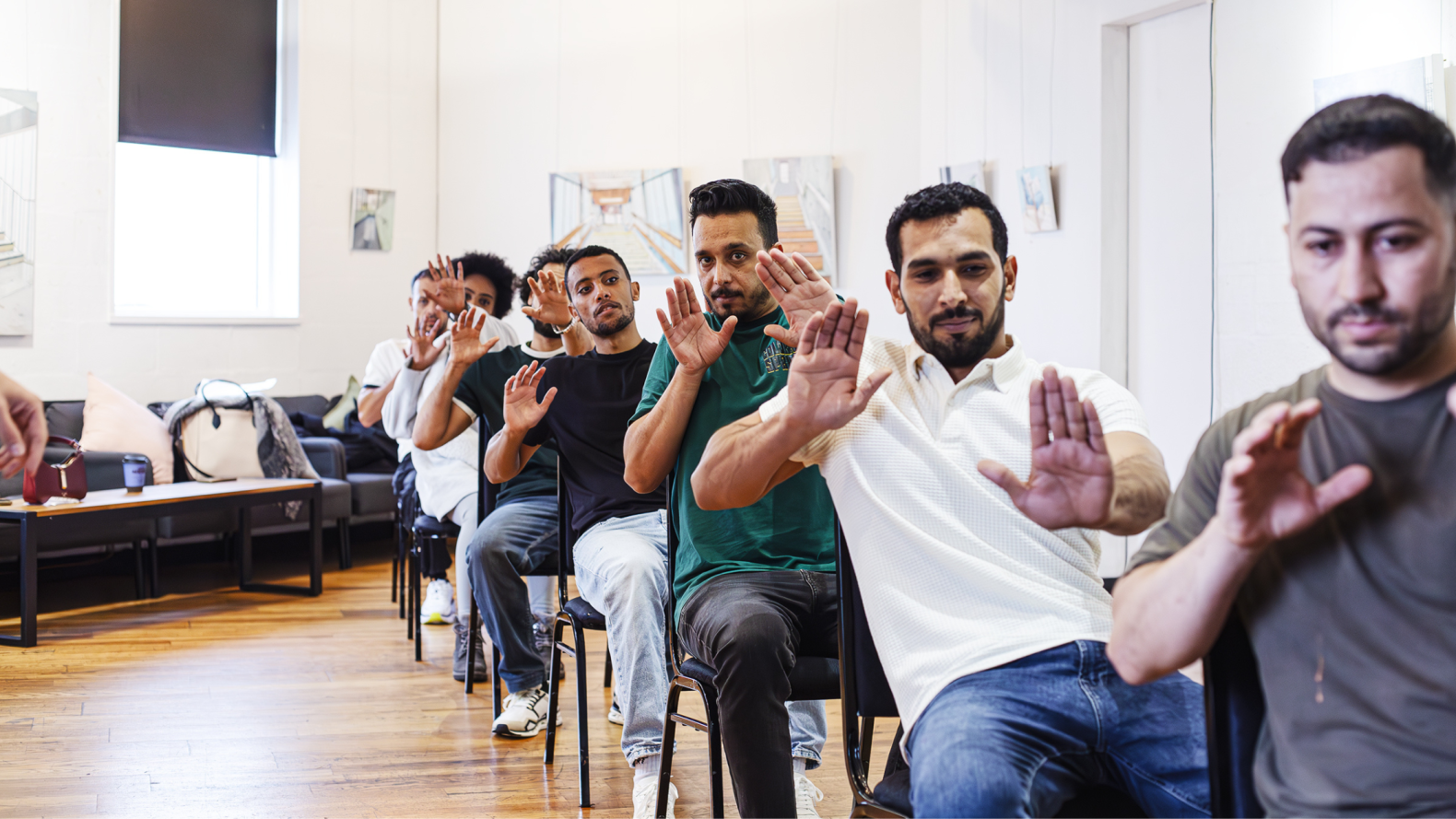 Seven people sitting on chairs, placed behind one another. All of them have both their hands out.
